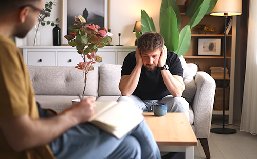 Ein Mann mit erschöpftem Gesichtsausdruck sitzt auf einem Sofa und stützt seinen Kopf in seinen Händen ab. 